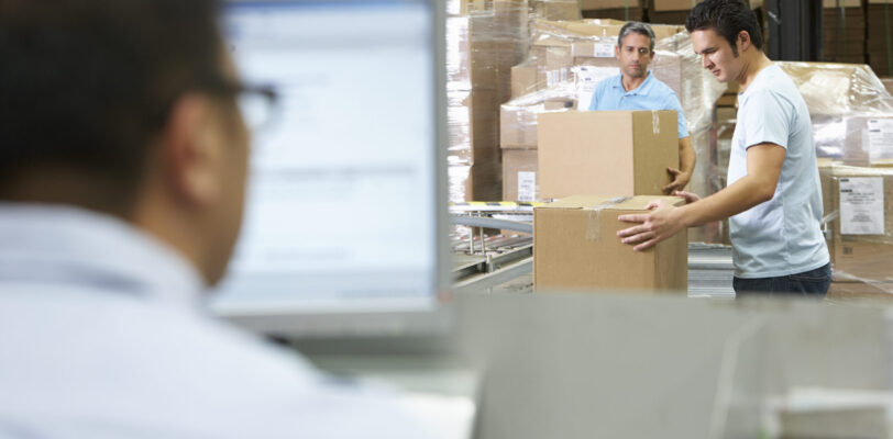 Warehouse workers package orders in boxes while a man uses an ERP on the computer.