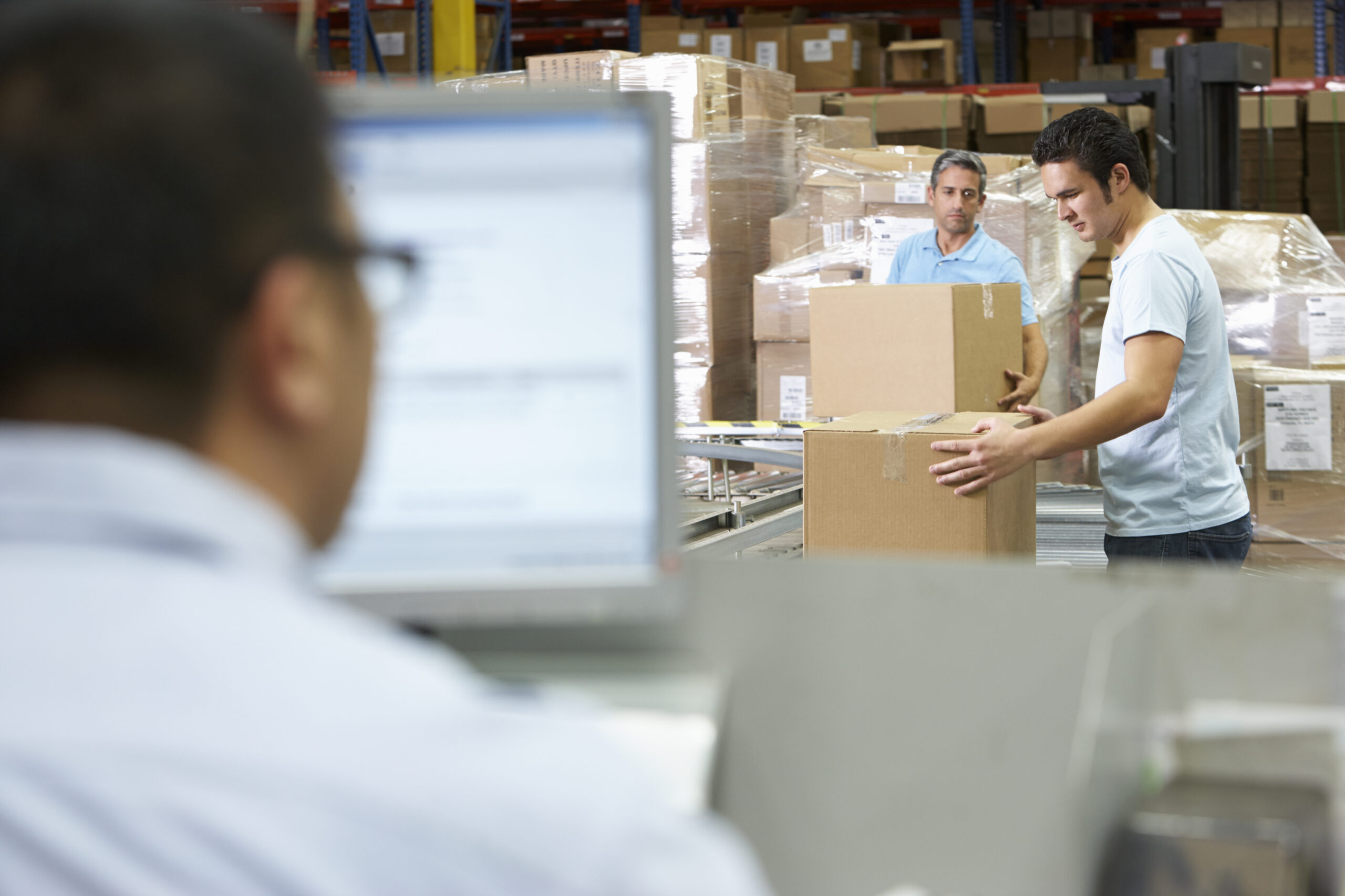Warehouse workers package orders in boxes while a man uses an ERP on the computer. 