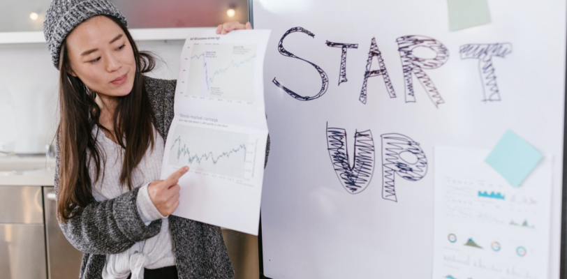 a woman gives a presentation while a poster next to her reads "Start Up"
